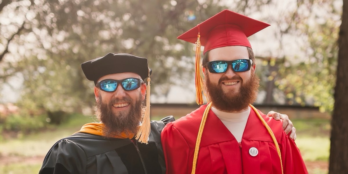 Joseph England, right, in a red cap and gown, standing with his community college math professor, left, in black and gold doctoral regalia, at graduation.